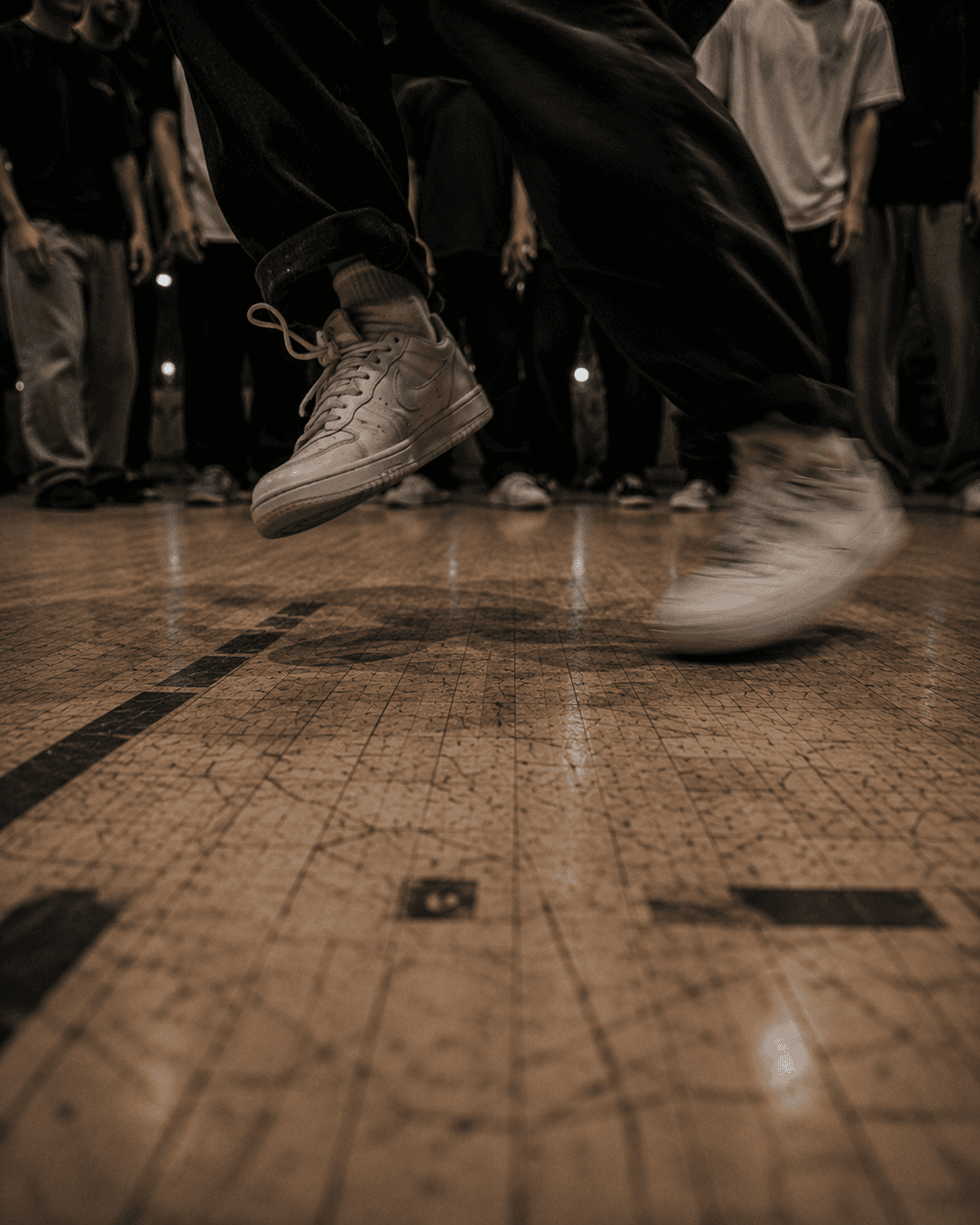 House footwork at full tempo, a dancer's feet caught mid-step above the scuffed studio floor while the circle watches from the edge of frame.